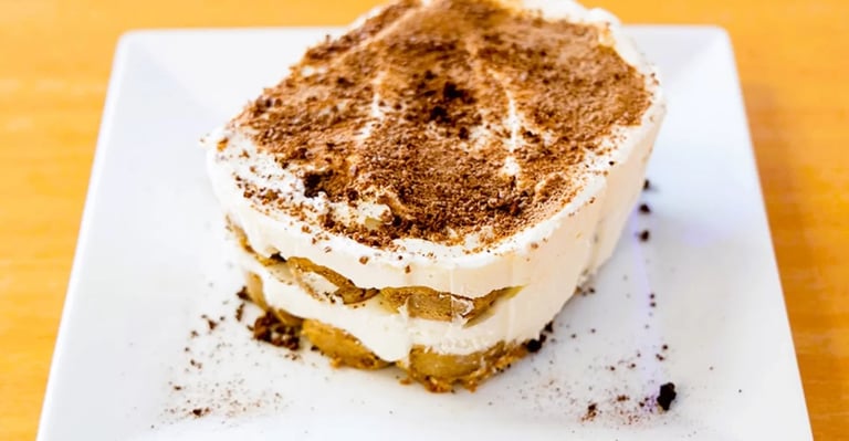 Square slice of tiramisu with cocoa powder topping on a white plate against a yellow background