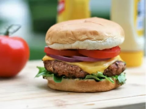 A stacked cheeseburger with tomato, lettuce, and cheese on a bun, with a tomato and beverages blurred in the background