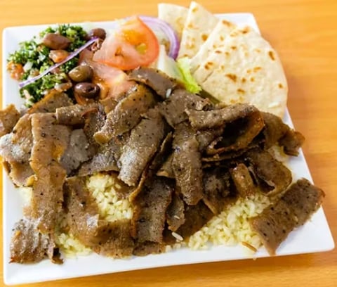 Plate of shawarma meat over rice with hummus, fresh vegetables, olives, and flatbread on the side