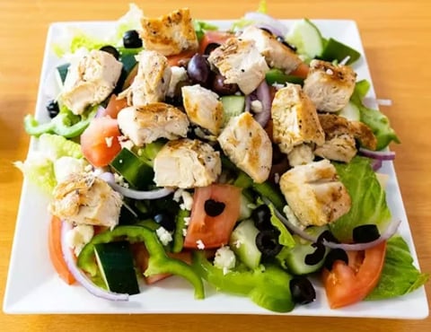 Grilled chicken salad with fresh greens, tomatoes, cucumbers, black olives, and crumbled cheese on a white square plate