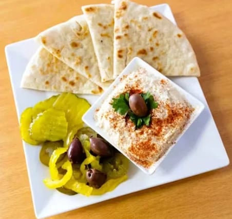 White plate with hummus topped with paprika and chickpea, flatbread triangles, and marinated olives with pickled peppers